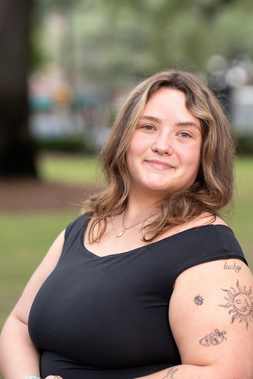 A person with shoulder-length brown hair and visible tattoos on their arm stands outdoors in a grassy park, smiling gently at the camera.