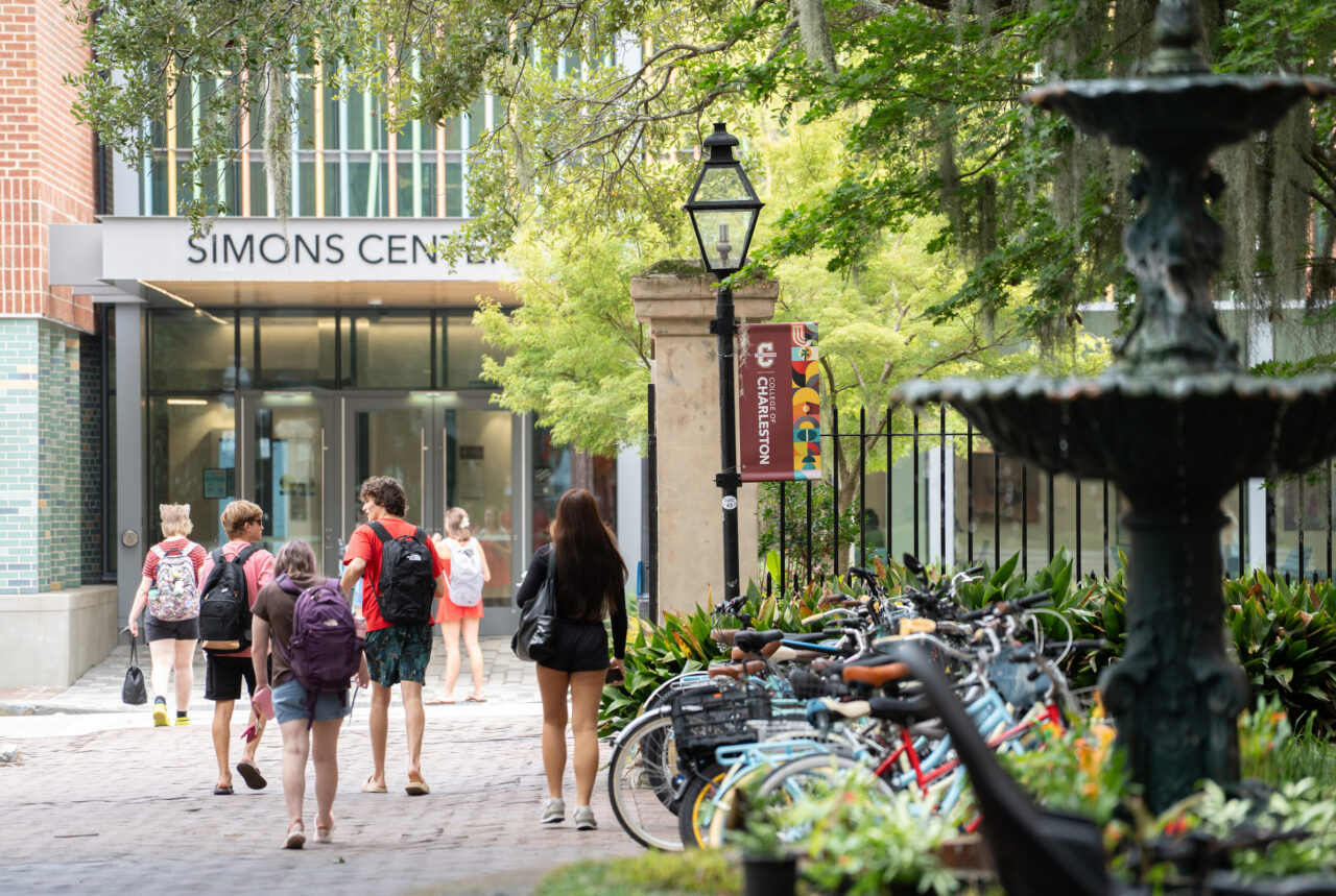 Students walk along a tree-lined path near the Simons Center, with bicycles parked nearby and a fountain in the foreground.