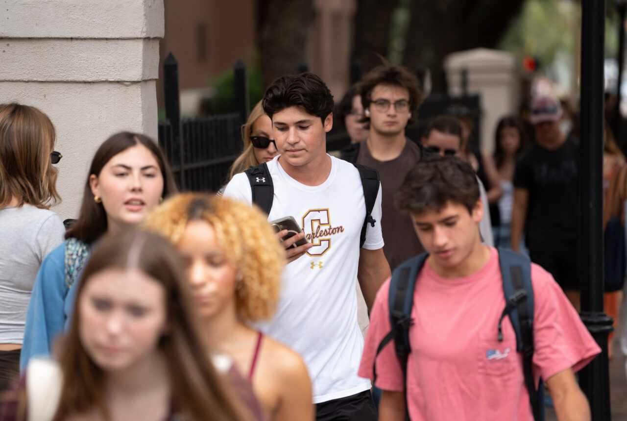 A group of young people walk along a city sidewalk, with one person in a white shirt featuring a "Carleton" logo looking at their phone.