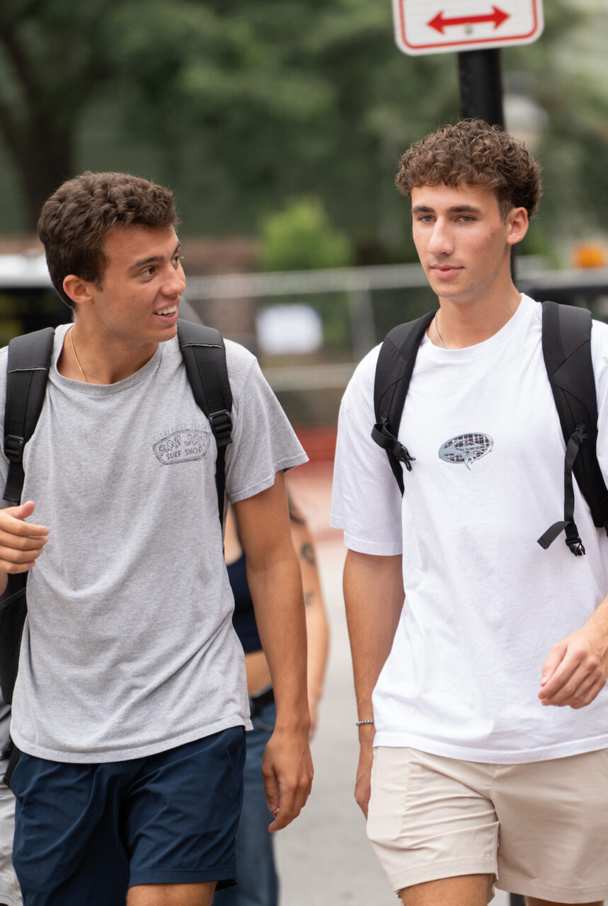 A group of young people walk together on a sidewalk, some carrying backpacks and holding water bottles, with trees and a street sign visible in the background.