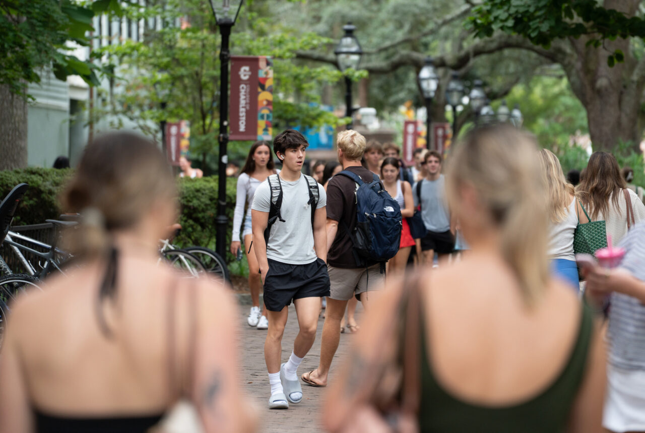 A group of people walk along a tree-lined path on a campus, with some carrying backpacks and others holding drinks.
