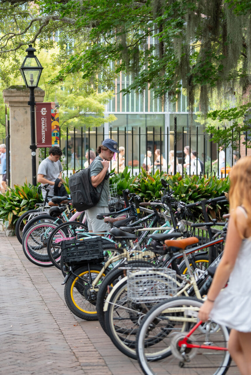 A person with a backpack stands among a row of bicycles on a brick-paved path under leafy trees, near a sign for the College of Charleston.