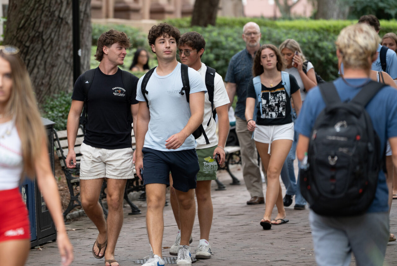A group of young people walks along a tree-lined sidewalk, with some carrying backpacks and others engaged in conversation.