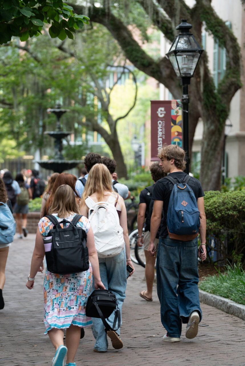 Students walk along a tree-lined brick pathway on a campus, carrying backpacks and bags.