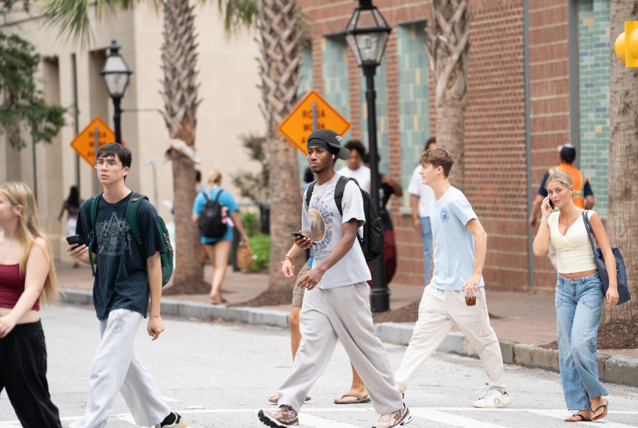 A group of young people cross a street in a sunny urban area with palm trees and brick buildings.