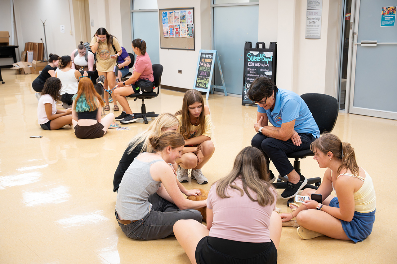 A group of young people sit in a circle on the floor in a brightly lit room, engaged in a collaborative activity with a facilitator seated nearby.