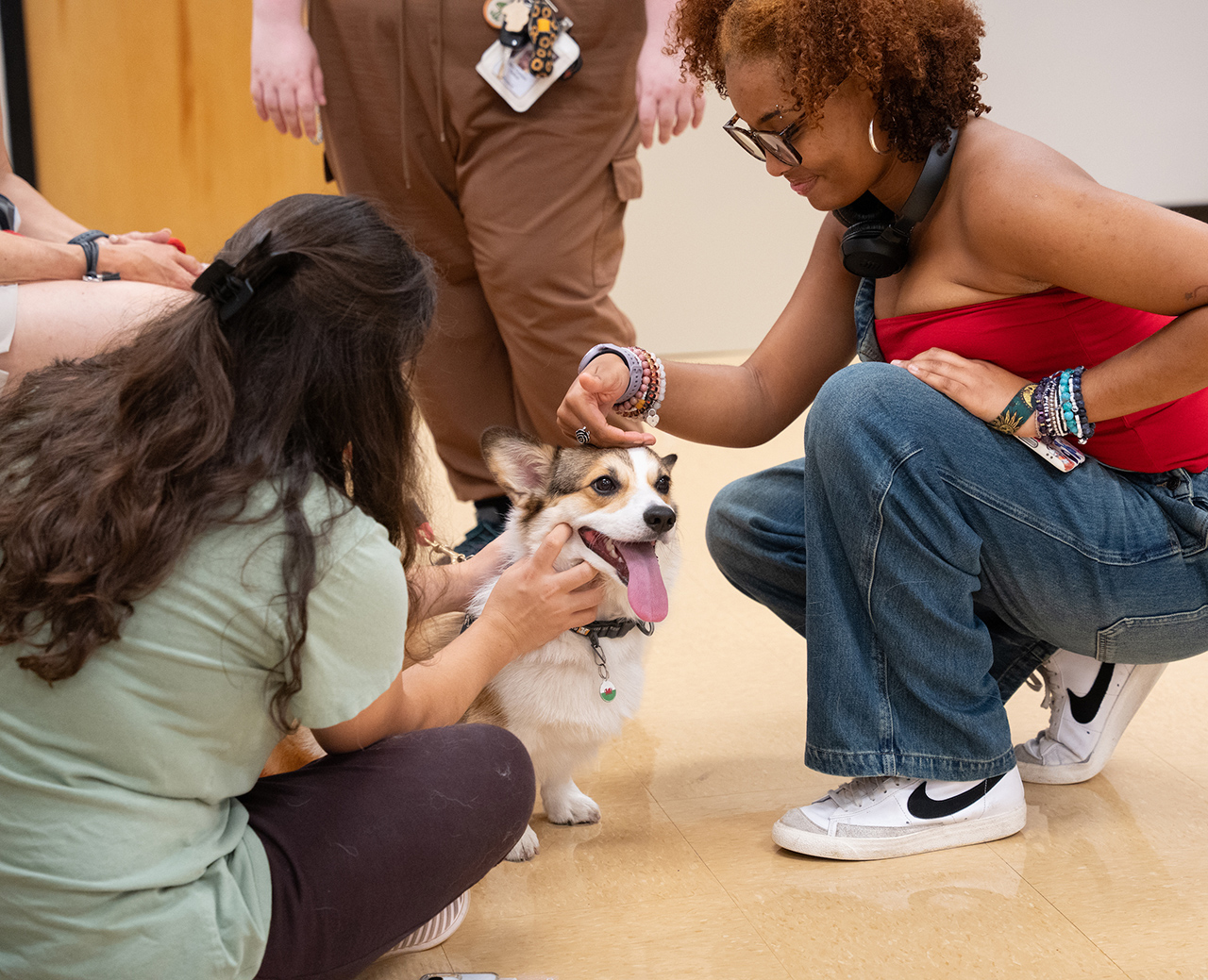 Two individuals are gently petting a happy corgi puppy while crouching on a tiled floor.