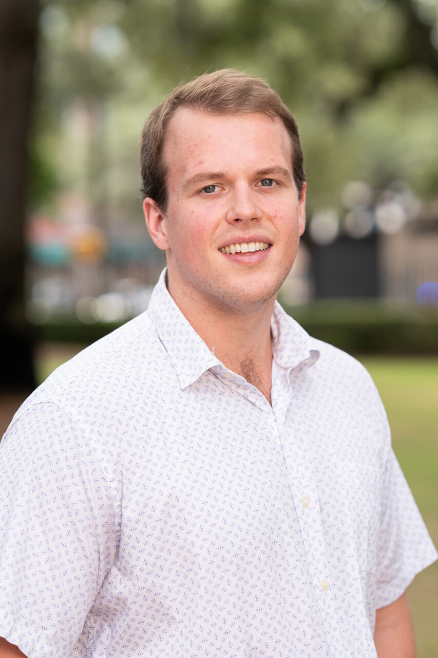 A person with short brown hair and a light complexion smiles while standing outdoors in a park-like setting, wearing a short-sleeved, patterned button-up shirt.