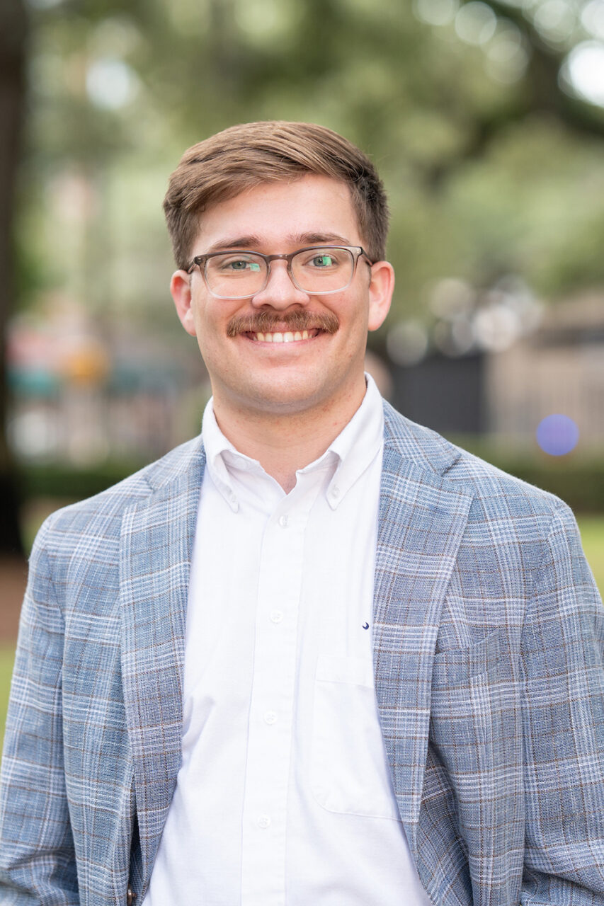 A person with glasses and a mustache smiles while wearing a plaid blazer over a white shirt, standing outdoors in a grassy area with trees in the background.