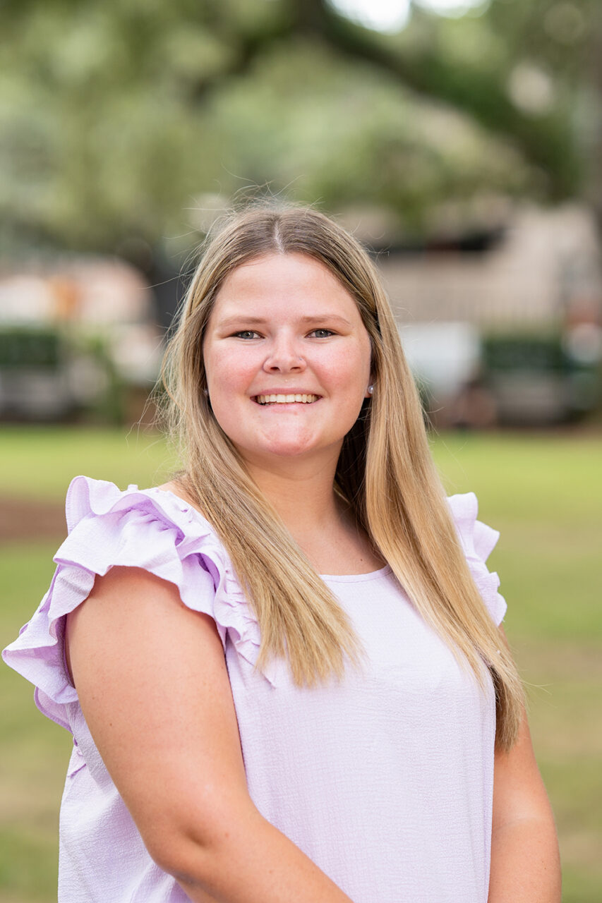 A young person with long blonde hair smiles at the camera while wearing a light purple top with ruffled sleeves, standing outdoors in a grassy area with trees in the background.