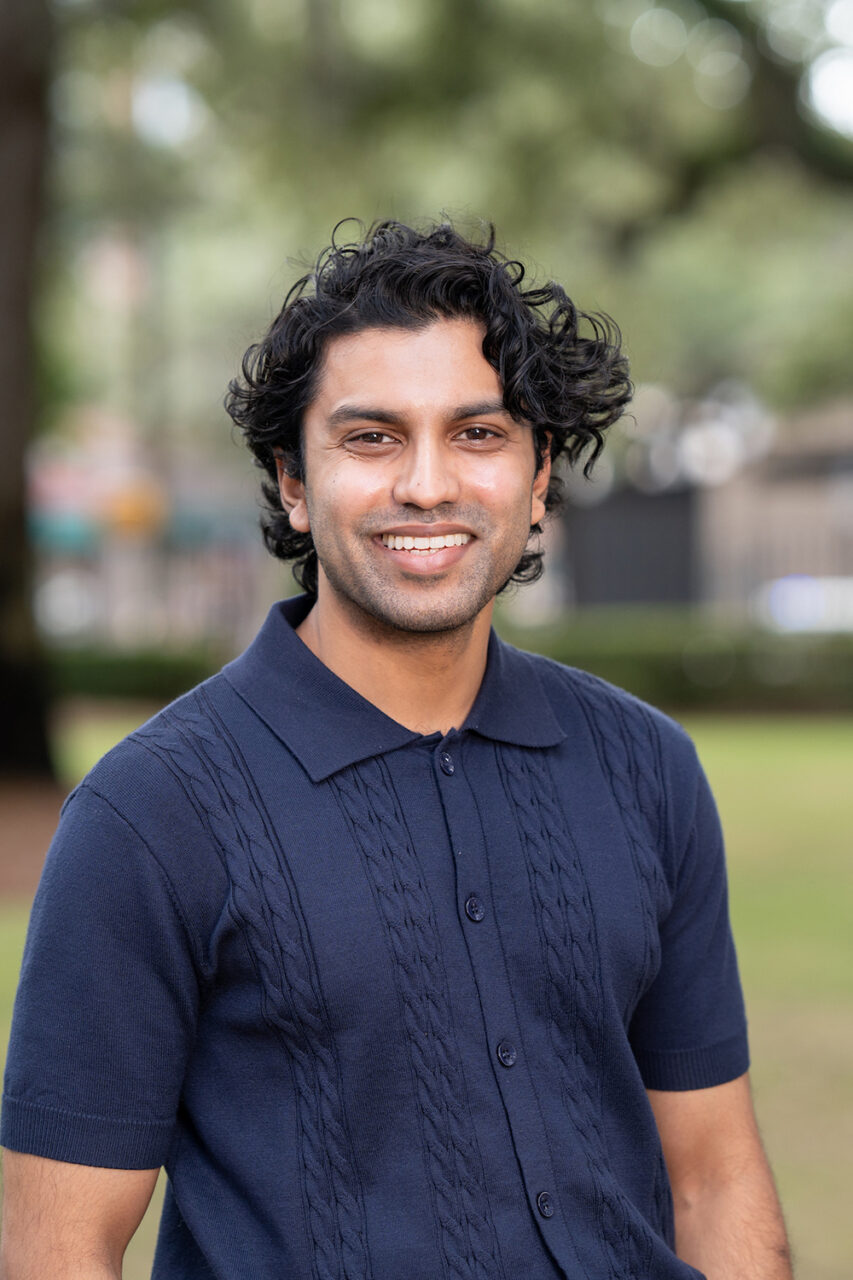 A person with curly dark hair smiles while standing outdoors in a grassy area with trees and blurred background elements.
