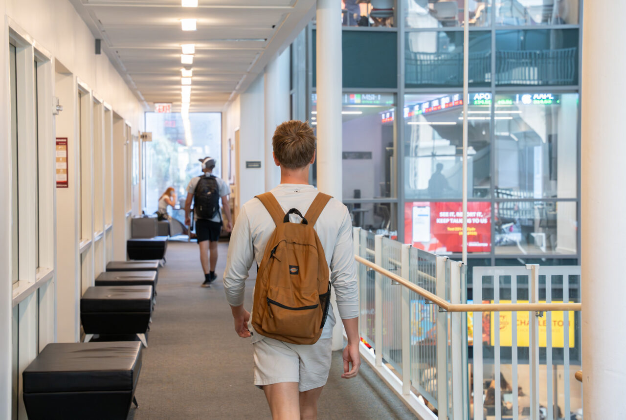 A person with a backpack walks down a modern hallway toward a glass-enclosed area, with others visible in the distance.