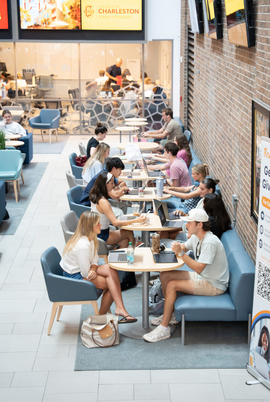 Students study and collaborate at round tables in a modern campus lounge with brick walls and digital displays.