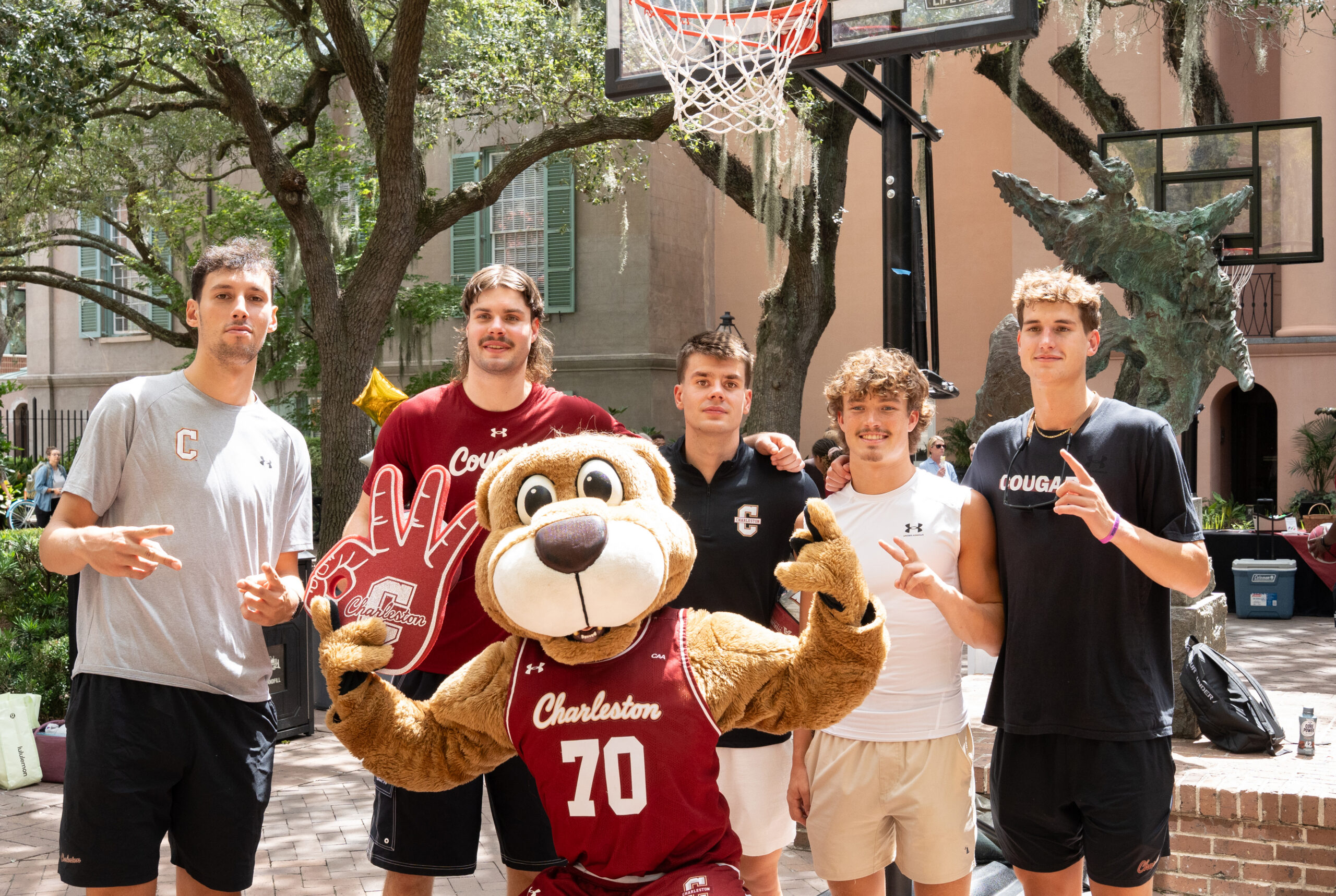 A group of young people, including a mascot wearing a Charleston jersey, pose together outdoors near a basketball hoop.
