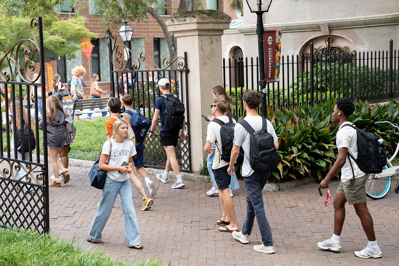 Students walk across a brick pathway near ornate gates and greenery at a university campus.