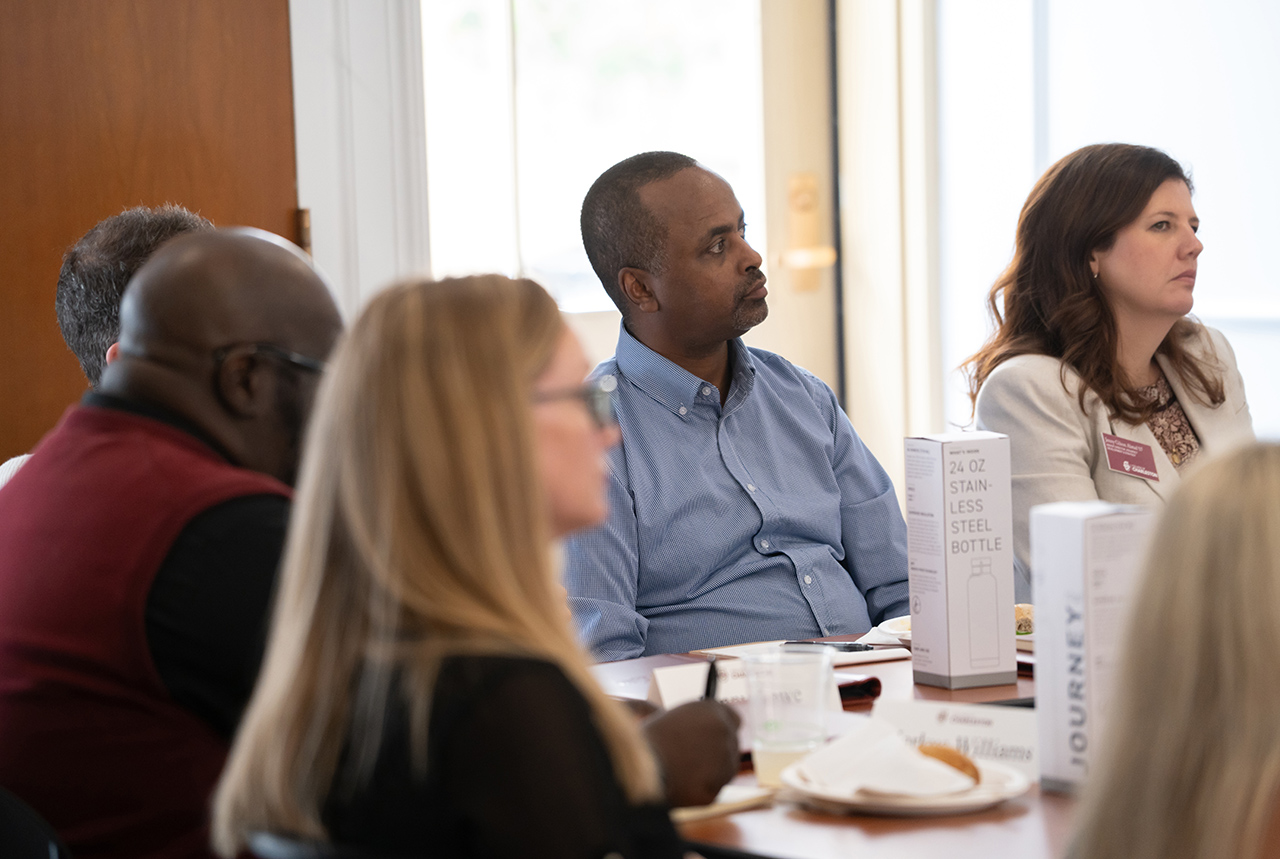 A group of people are seated at a table in a meeting, with one individual in a blue shirt looking attentively toward the side.