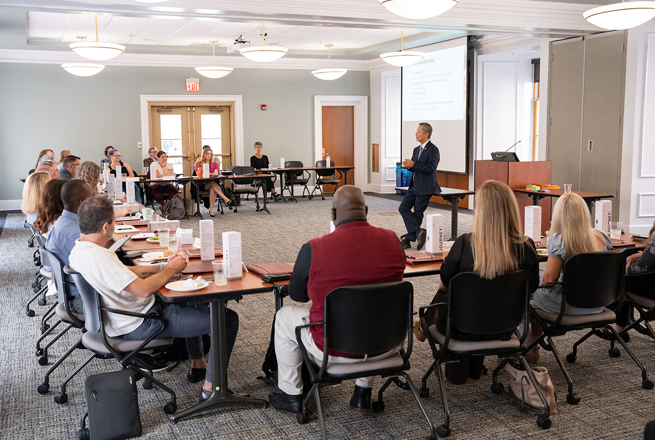A person stands at a podium presenting to an audience seated at tables in a conference room.
