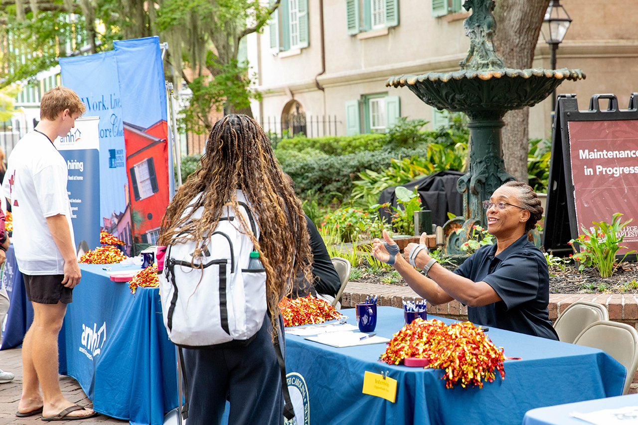 A person with long dreadlocks engages with a smiling representative at a blue-clothed table during an outdoor event, with another individual nearby and a fountain in the background.