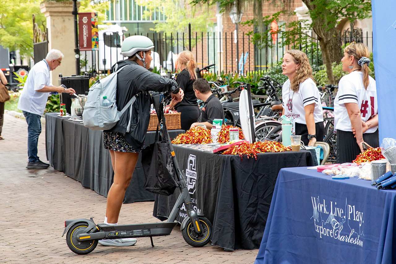 A person on an electric scooter interacts with a table displaying promotional items at an outdoor event in Charleston.