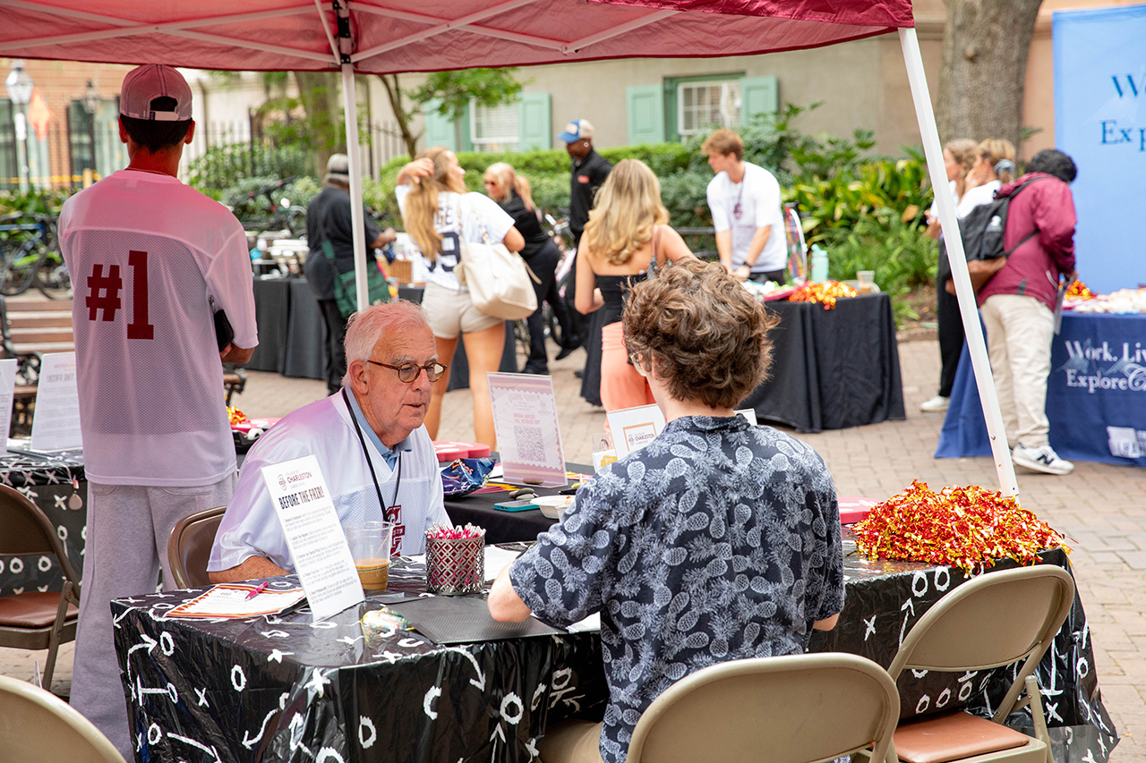 A person in a #1 jersey stands near a table under a red canopy where another individual is seated, engaging with a vendor at an outdoor event.