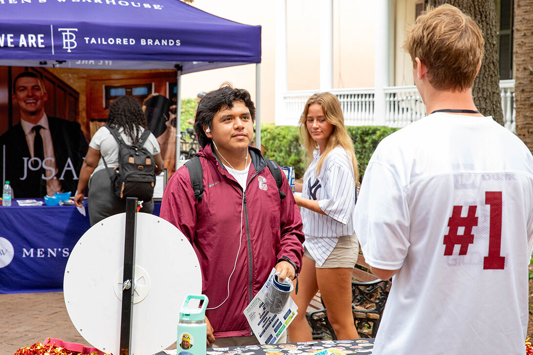 A person in a maroon jacket stands at a Men's Wearhouse booth, engaging with another individual wearing a white jersey with "#1" on the back, while others browse nearby.