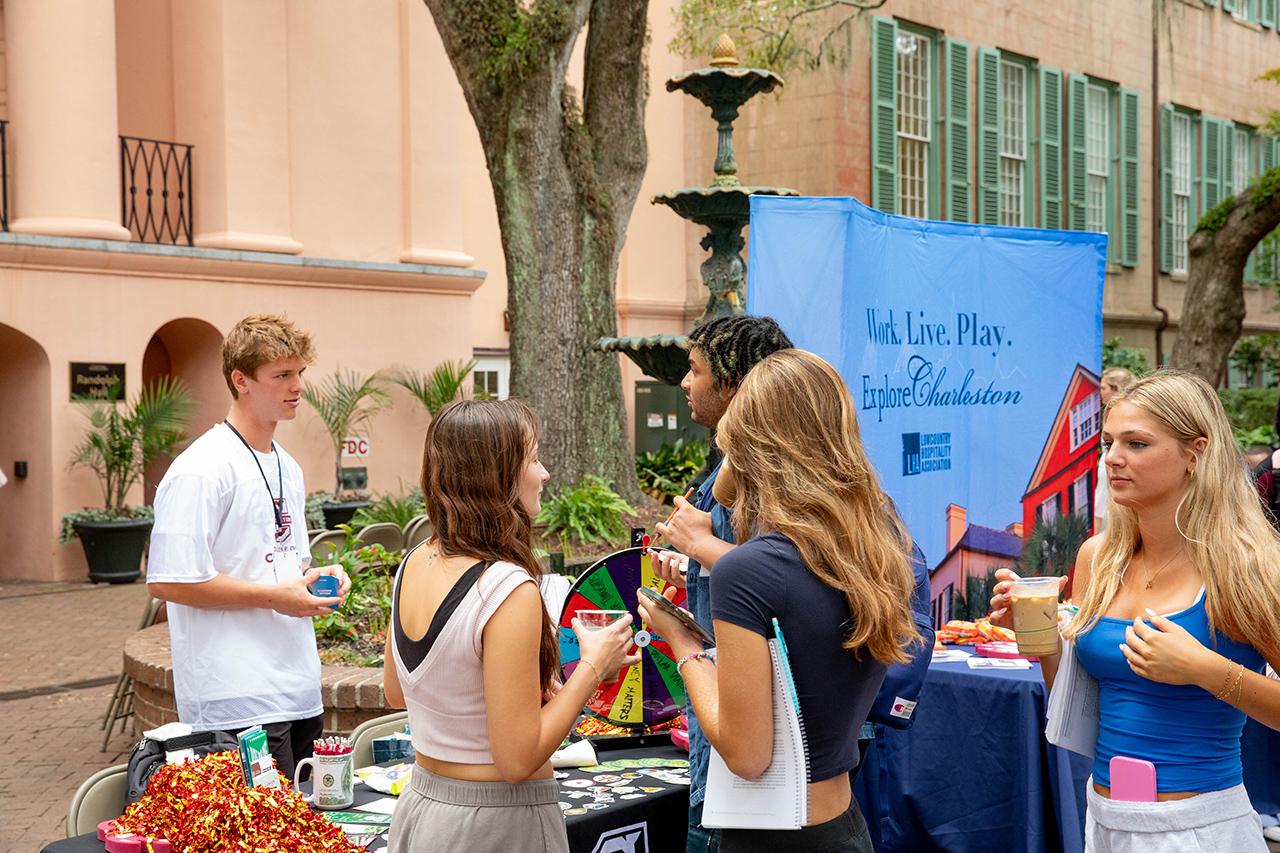 A group gathers outdoors near a fountain and a "Work. Live. Play. Explore Charleston" banner, engaging with a colorful wheel and refreshments at a promotional booth.