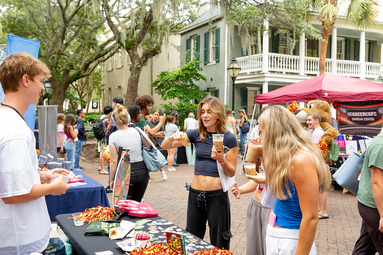 A group of young people gather outdoors at a campus event, interacting with vendors and sampling food and drinks under trees and historic buildings.