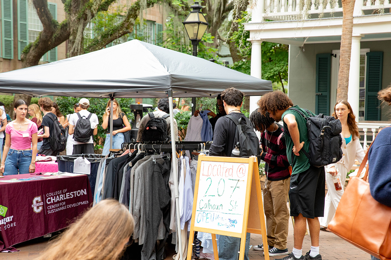 A group of people gather around a market stall under a canopy, browsing clothing and interacting with vendors at an outdoor event.