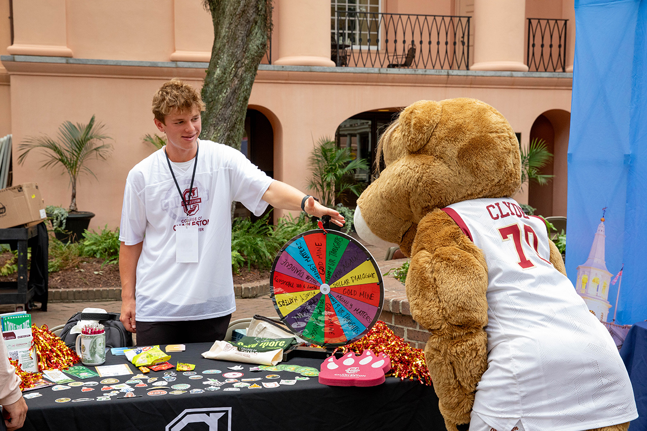 A person in a white shirt interacts with a large brown mascot wearing a jersey marked "CLYDE 70" at an outdoor event booth featuring a colorful wheel and various merchandise.