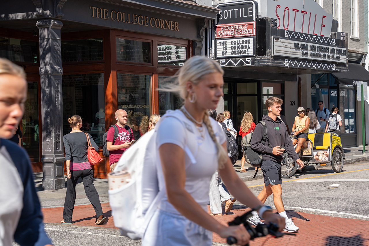 People walk and ride bicycles along a sunny street in front of storefronts including