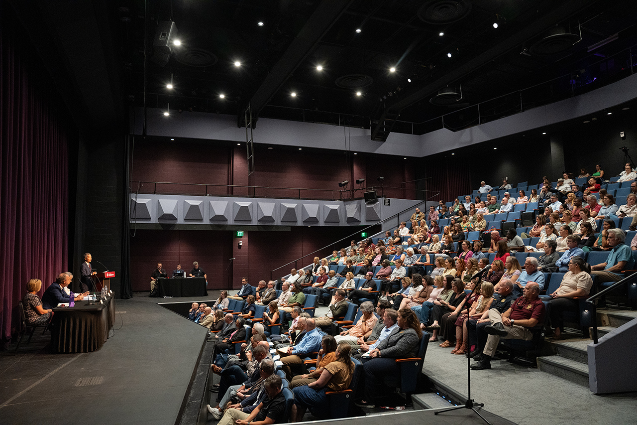 A panel discussion is taking place in a modern auditorium with an audience seated in tiered rows, attentively listening to speakers on stage.