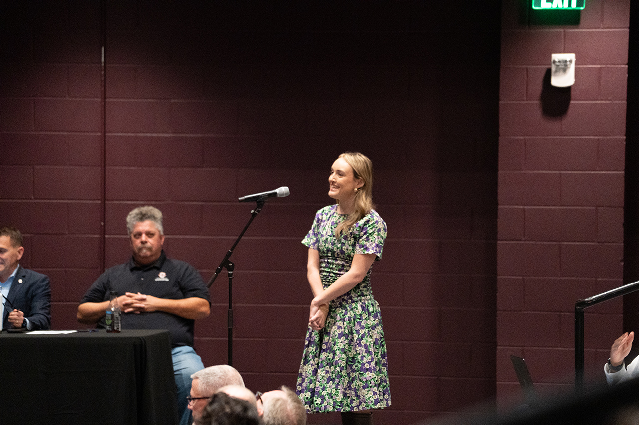 A person in a floral dress stands at a microphone on a stage, smiling, while others sit at a table behind them.