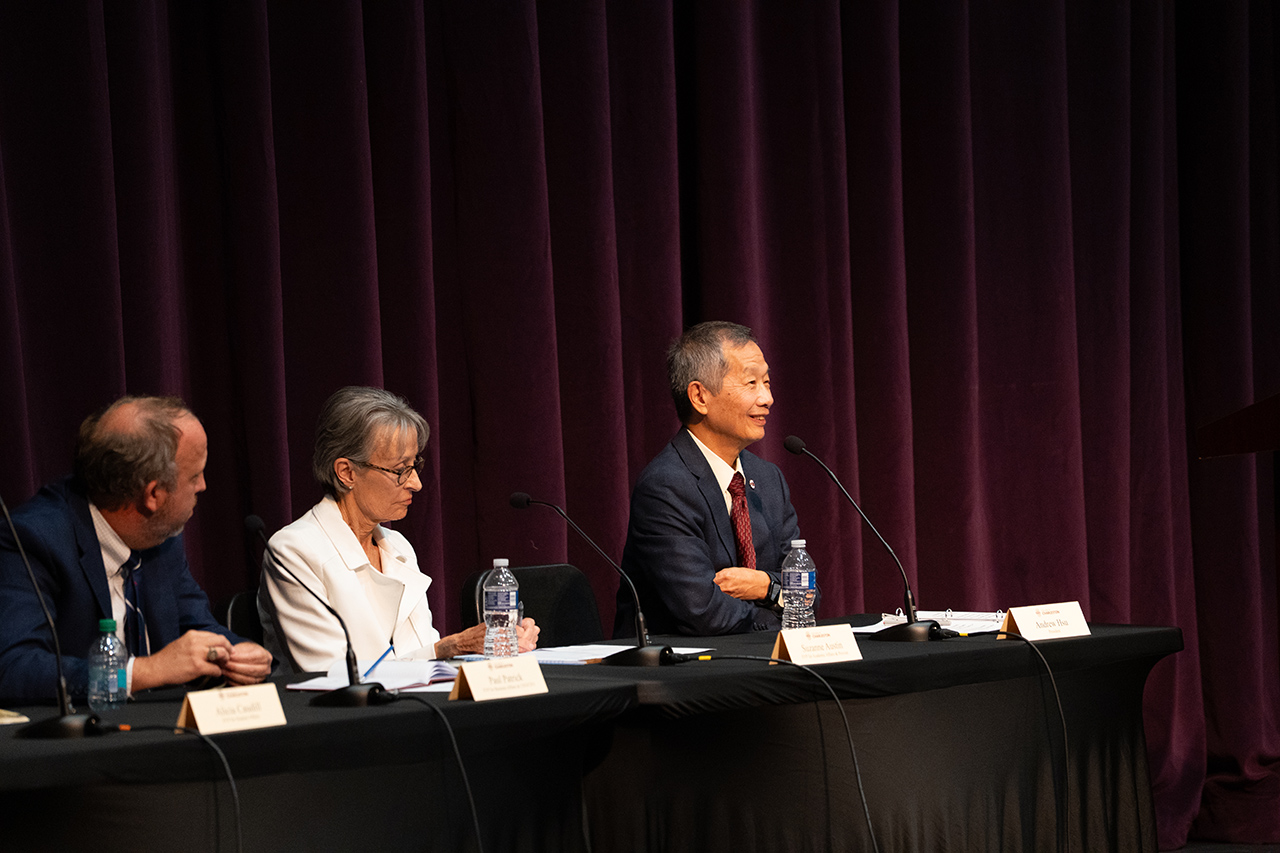 Three individuals sit at a long table with microphones and nameplates, facing an audience against a dark curtain backdrop.