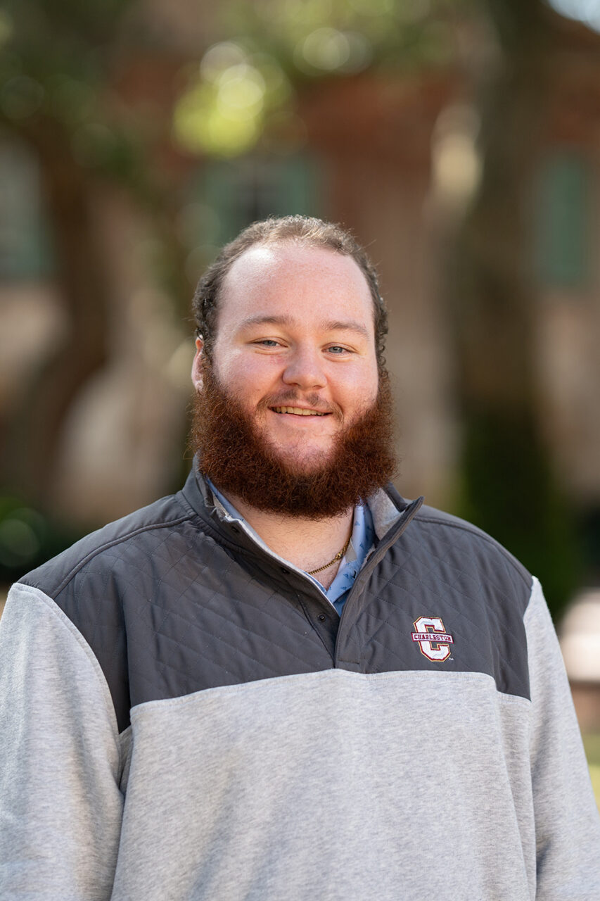 A person with a full beard smiles while wearing a gray and dark gray pullover with a South Carolina Gamecocks logo, standing outdoors in front of a blurred background of trees and buildings.