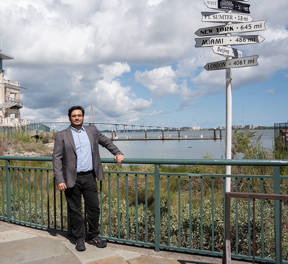 A person in a suit stands beside a railing near a waterfront, with a signpost indicating distances to various cities and landmarks, and a bridge visible in the background.