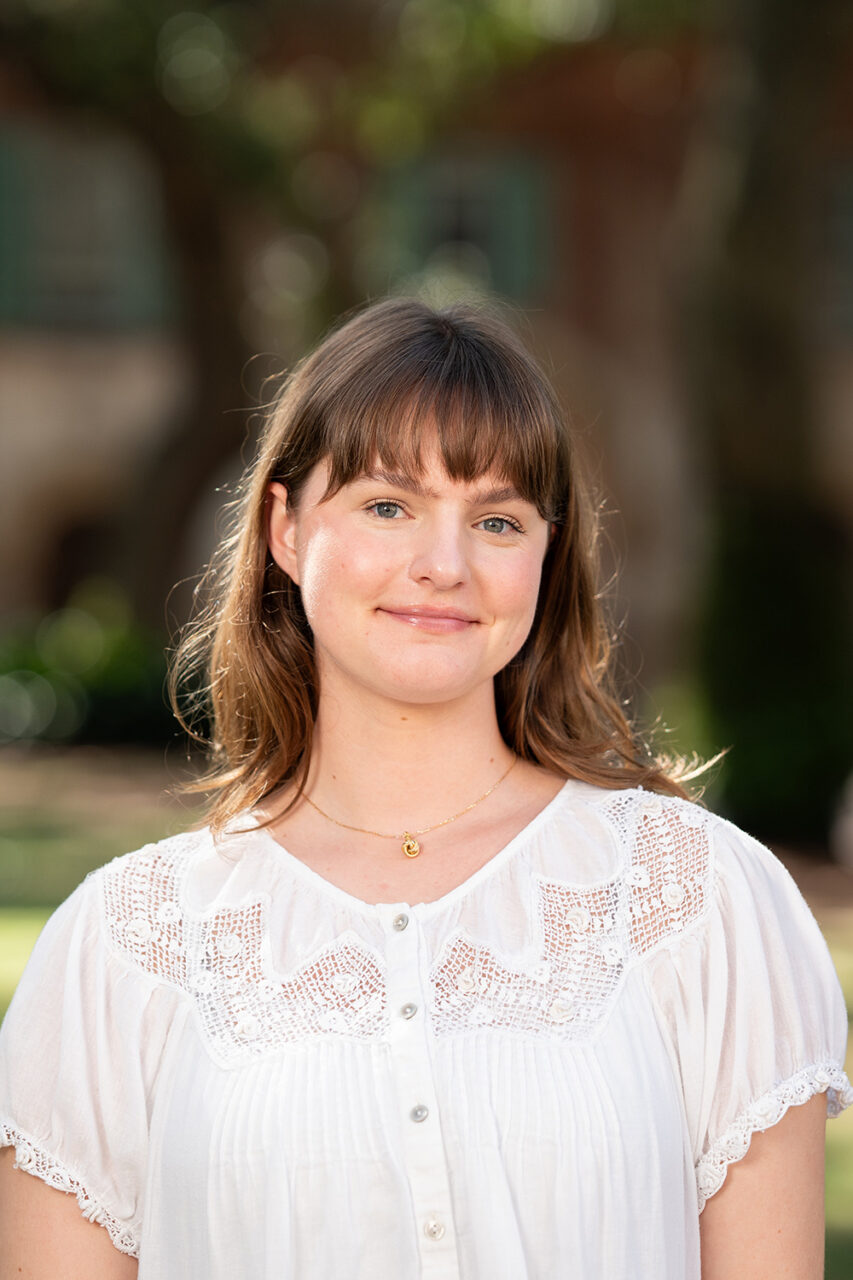 A young person with brown hair and bangs smiles gently while wearing a white blouse with lace details, standing outdoors in a sunlit park setting.