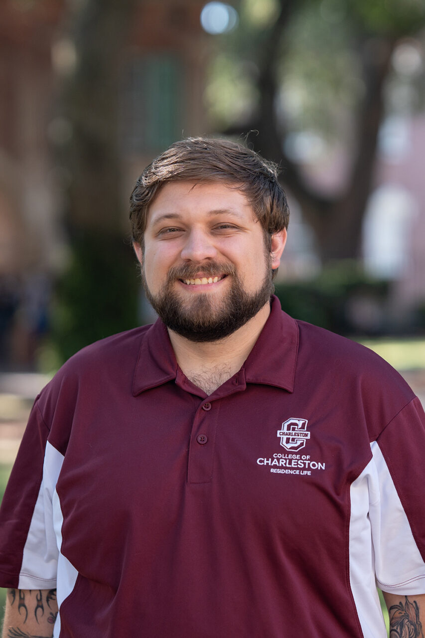 A person with a beard smiles while wearing a maroon polo shirt featuring the College of Charleston logo, standing outdoors on a sunny day with trees and buildings in the background.