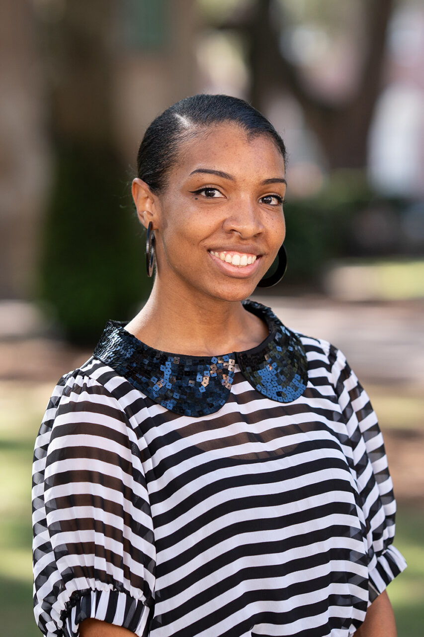 A smiling person stands outdoors in a park-like setting, wearing a black-and-white striped top with a decorative collar.