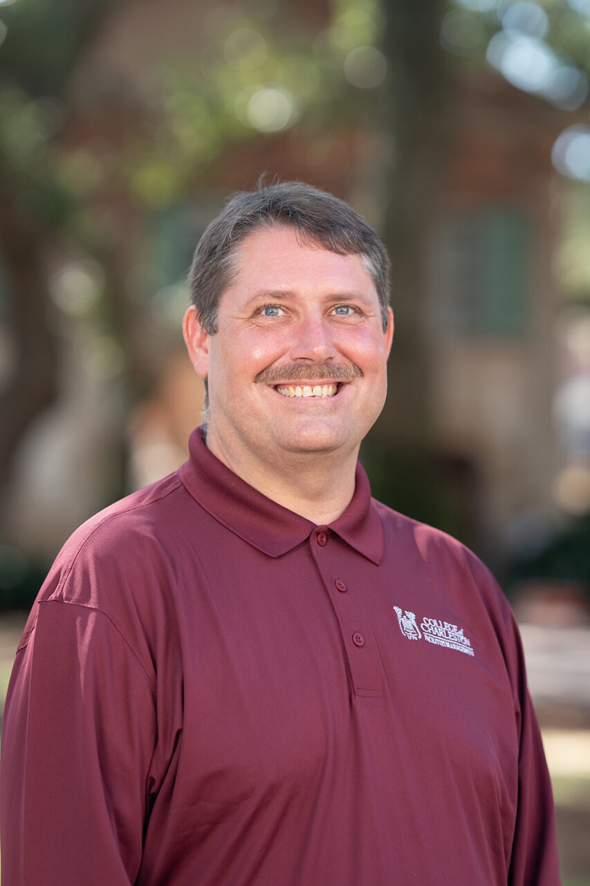 A person wearing a maroon polo shirt with a logo smiles outdoors in front of a blurred background of trees and buildings.
