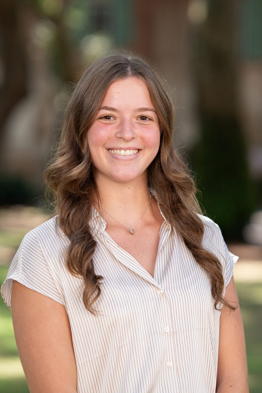A young person with long brown hair smiles warmly while standing outdoors in a sunlit, tree-shaded area.