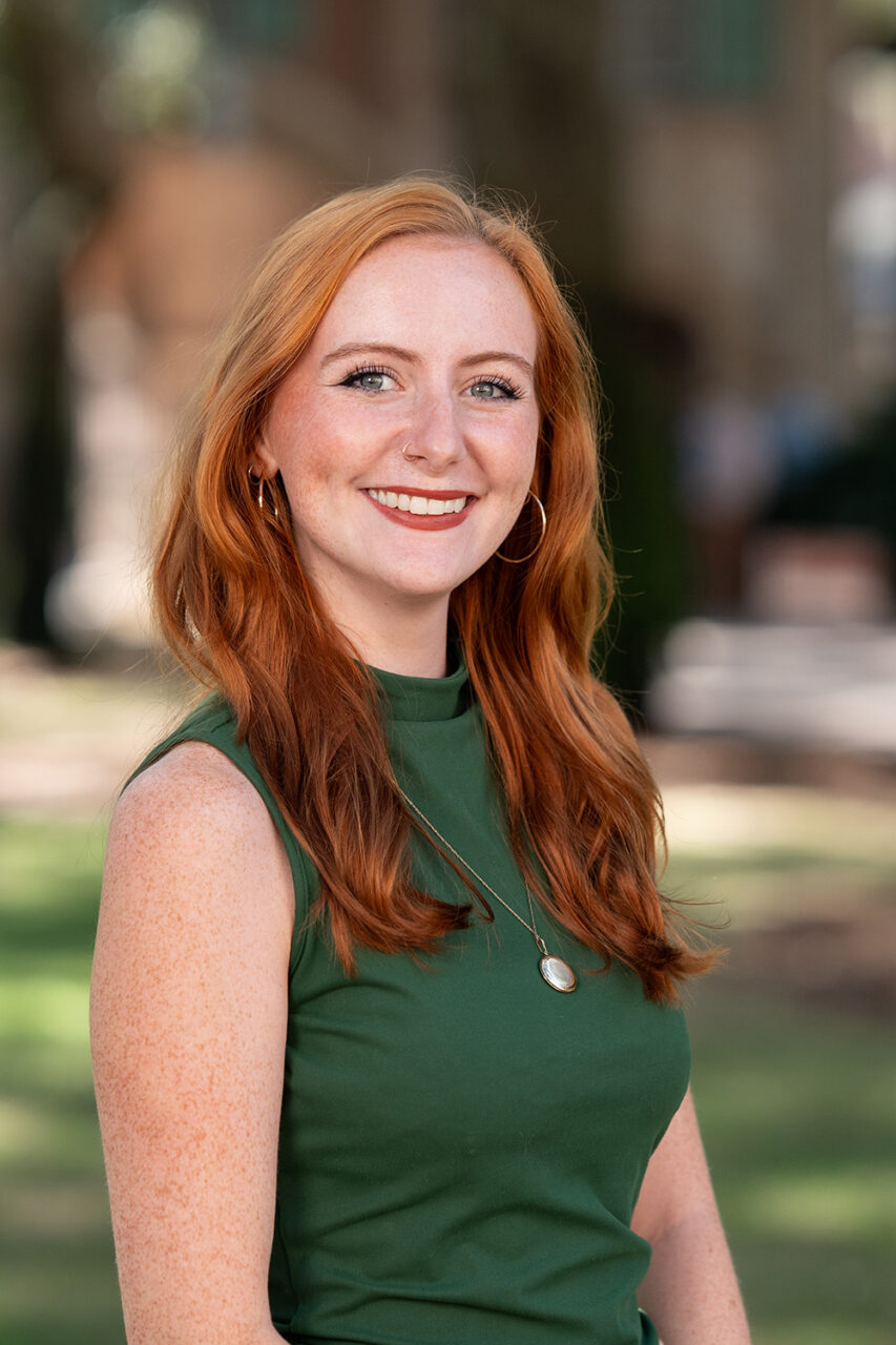 A person with long red hair smiles while wearing a green sleeveless top, standing outdoors in a sunlit park with trees and buildings in the background.