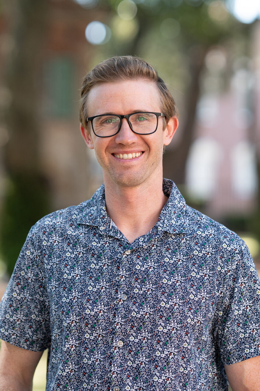 A person with glasses and a patterned shirt smiles outdoors in a sunlit, tree-lined area.