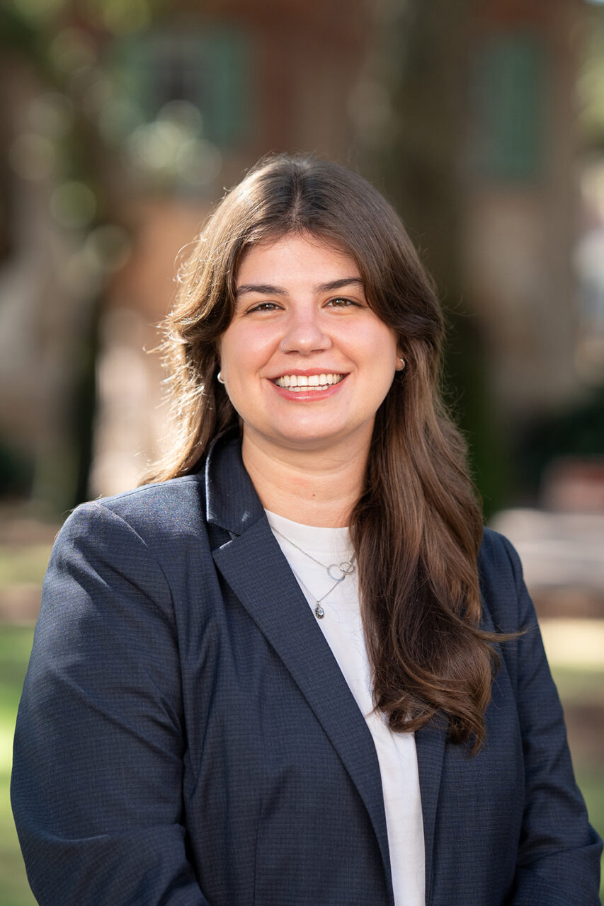 A smiling person in a dark blazer stands outdoors on a sunny day with trees and buildings in the background.