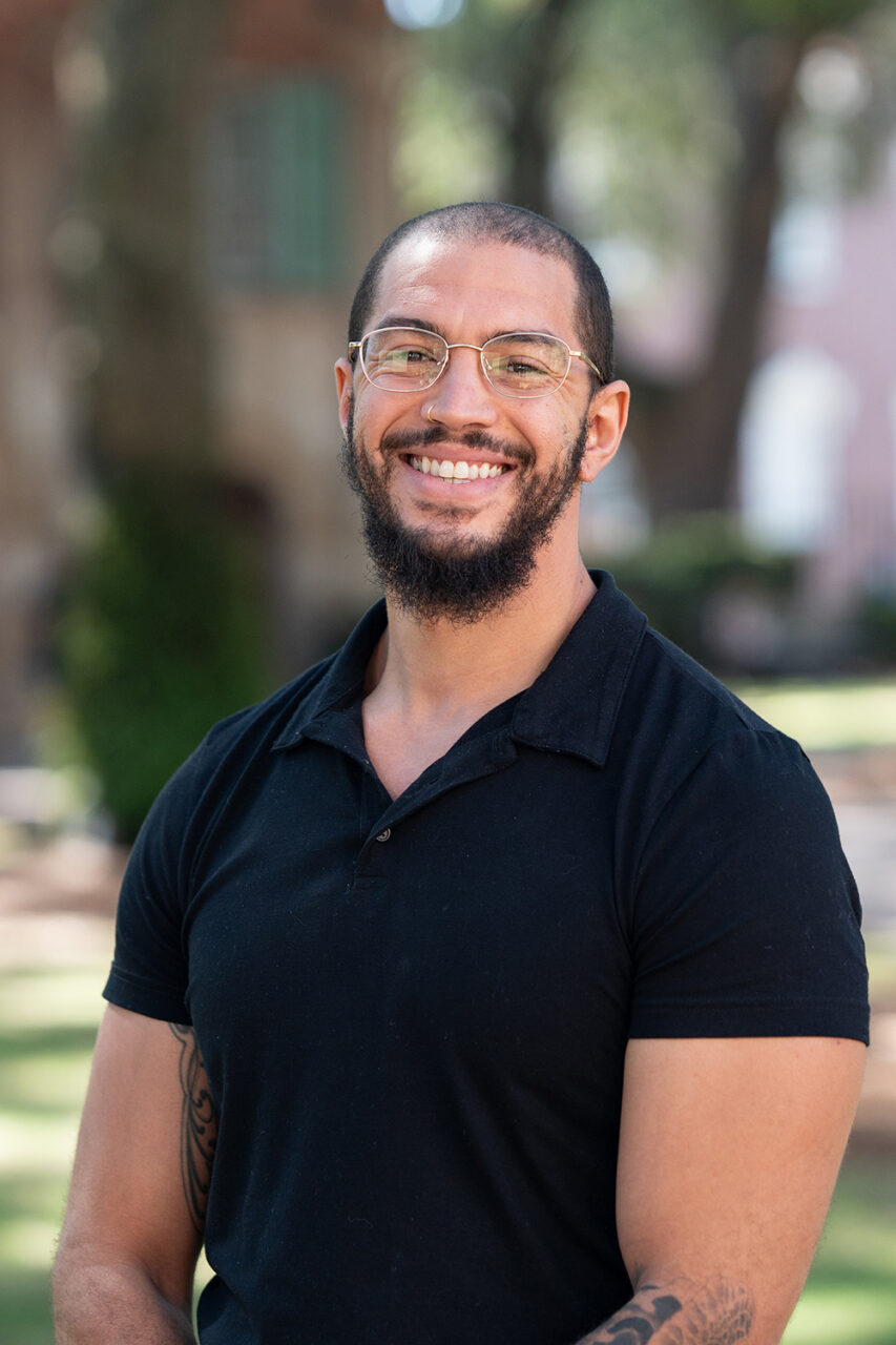 A smiling person with glasses and a beard stands outdoors in a park-like setting, wearing a black polo shirt.