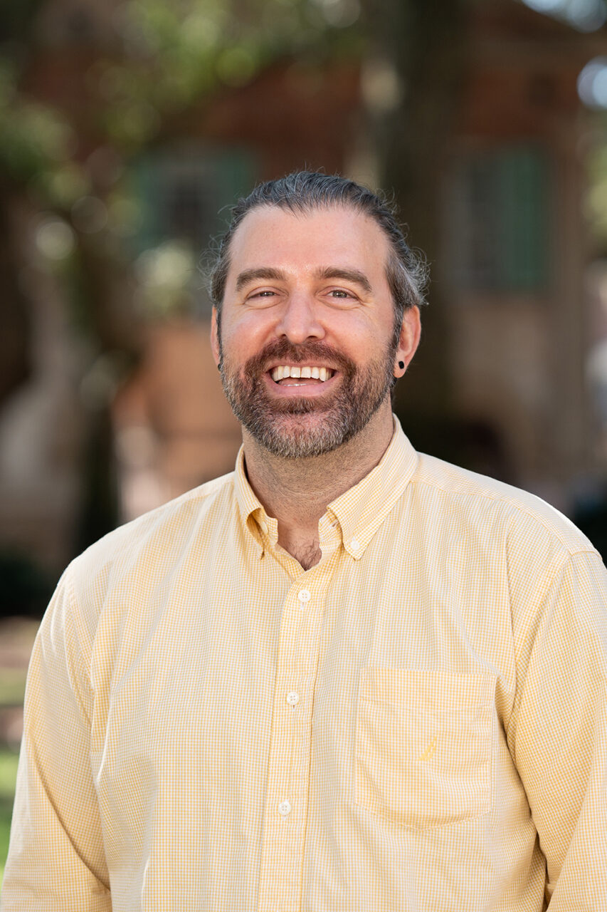 A smiling person with a beard stands outdoors in a sunlit area with trees and a building in the background.