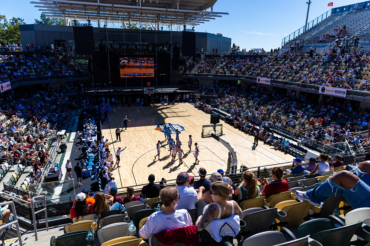A basketball game is underway in a large outdoor arena filled with spectators watching from tiered seating under a clear blue sky.