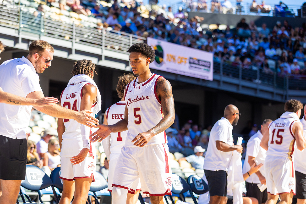 A basketball team in white and red uniforms gathers on the court during a game, with players and staff interacting near the sidelines as spectators watch from the stands.