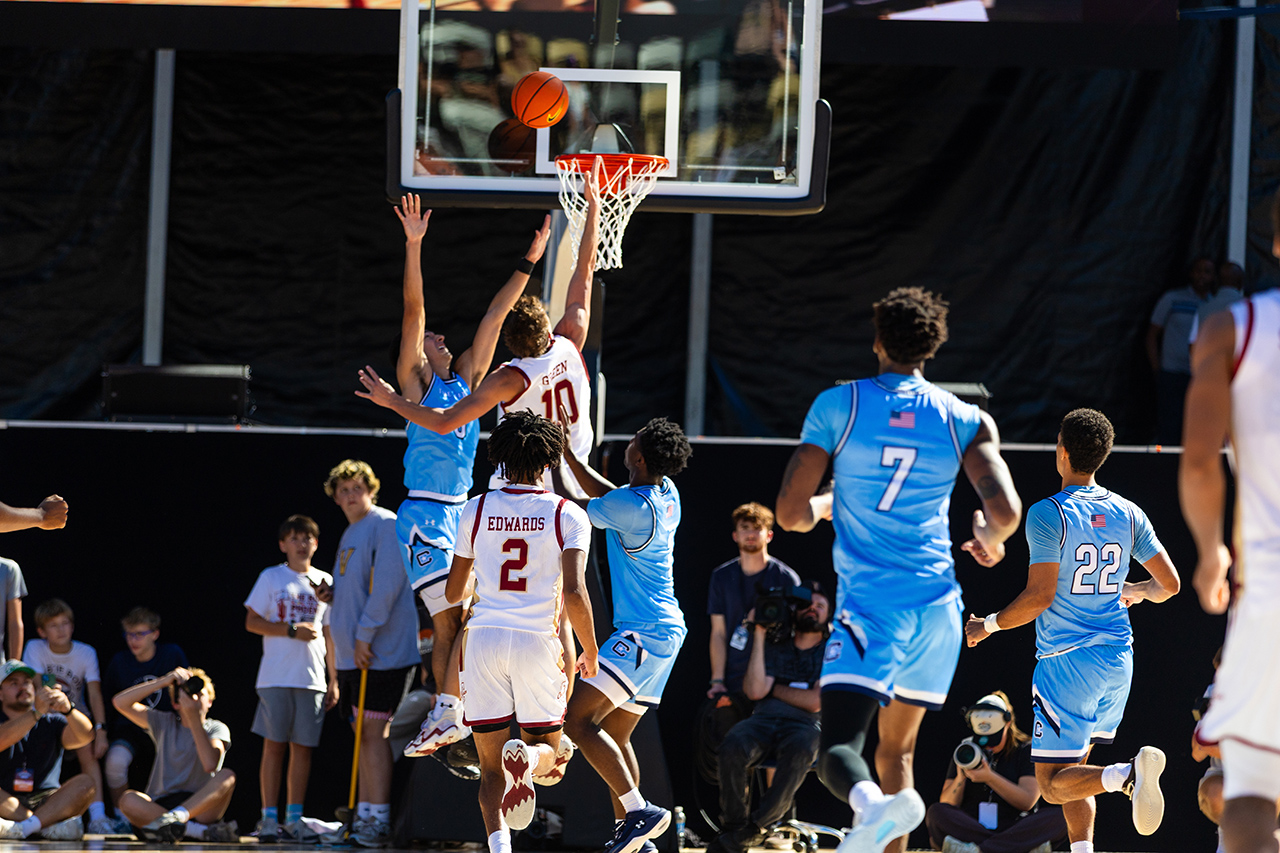A basketball player in a white jersey dunks the ball while being contested by opponents in blue uniforms during a game.