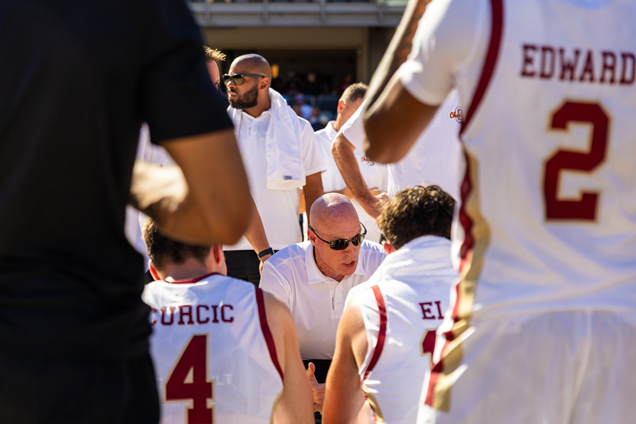 A coach in a white polo shirt and sunglasses speaks to players in a huddle during a basketball game, with jerseys displaying names and numbers.