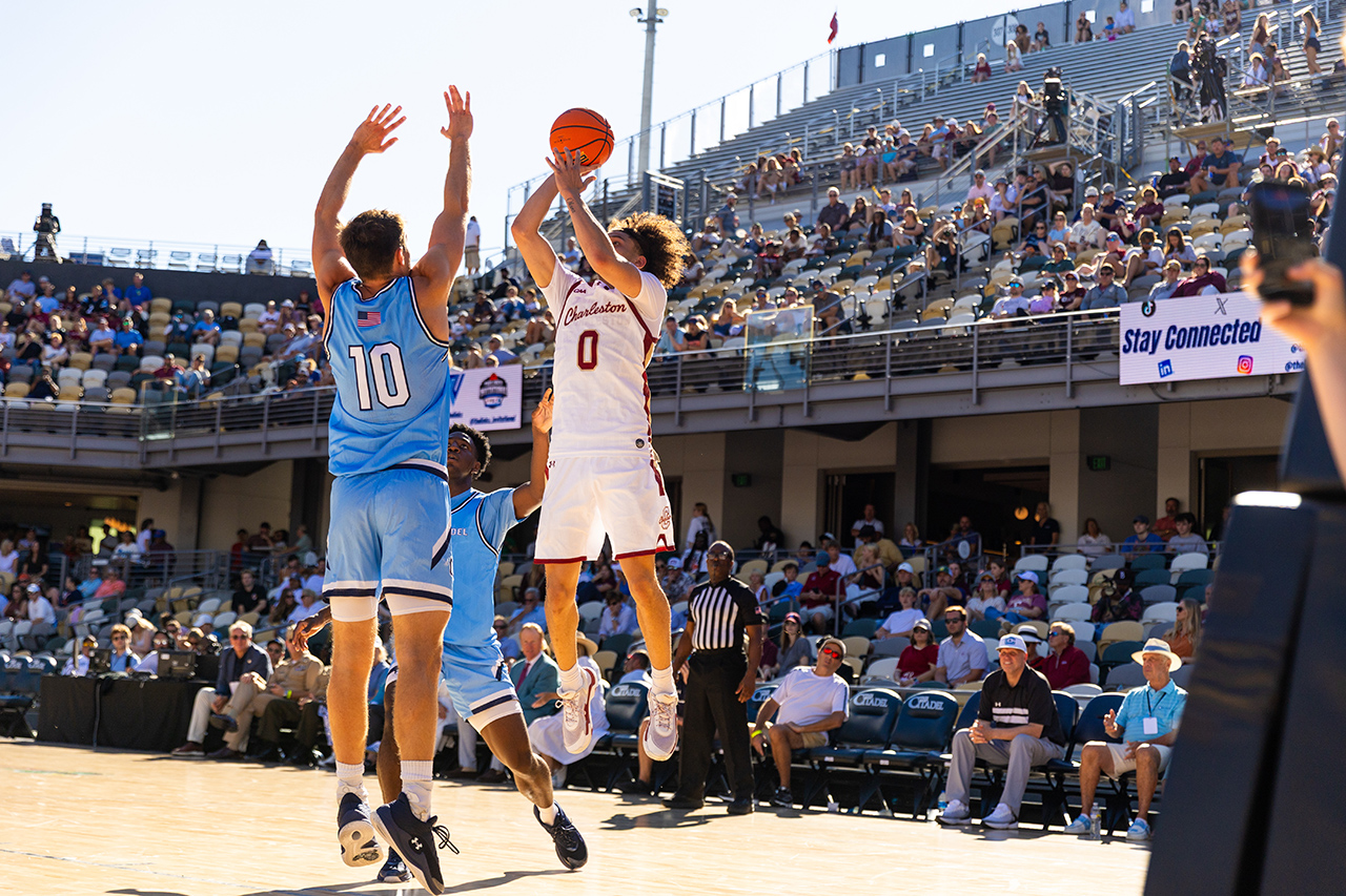 A basketball player in a white jersey with the number 0 leaps to shoot the ball while being defended by an opponent in a blue jersey with the number 10, during a game in front of a crowd.